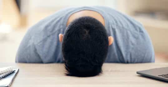Head down on the desk, wearing a gray shirt, they seem drained. A notebook and laptop flank them, hinting at tasks waiting to be tackled. Perhaps some motivation tips could lift their spirits when they just don't feel like doing anything.