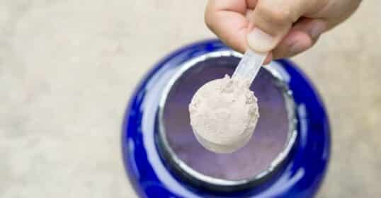 A hand holds a scoop of protein powder, hovering above an open blue container, with a hint of creatine ready to boost your workout mix.