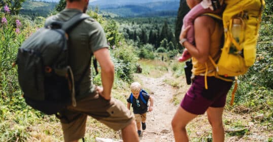 A family with two adults and two kids enjoys hiking on a forest trail. The child in the middle wears a blue backpack, while the adult on the right carries the other child.