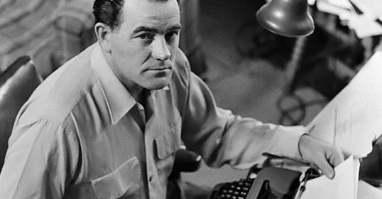 A person seated at a desk with a typewriter, immersed in channeling the legacy of Louis L'Amour, looks up at the camera as if ready to share a story fit for a podcast.