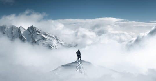A high-agency individual stands atop a snow-covered mountain peak, surrounded by clouds, with a majestic mountain range in the background, showcasing the characteristics of determination and resilience.