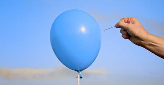 A hand holds a needle near a blue balloon against a clear sky, symbolizing the tension of rejection and how to deal with it.