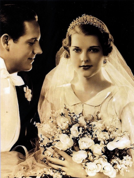 A black-and-white vintage wedding photo captures a moment in history, where the bride, with a bouquet and veil, stands beside her groom in a suit, symbolizing the timeless tradition of marriage.