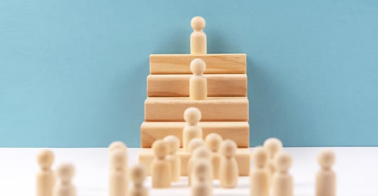Wooden figures are positioned in a pyramid shape on blocks, with a crowd of figures at the base, symbolizing the cult of leadership against a blue background.