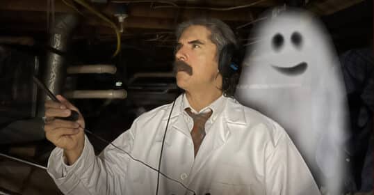 A man in a lab coat and headphones uses a device in a dimly lit room, capturing ghost voices. A cartoon-like spirit appears beside him.