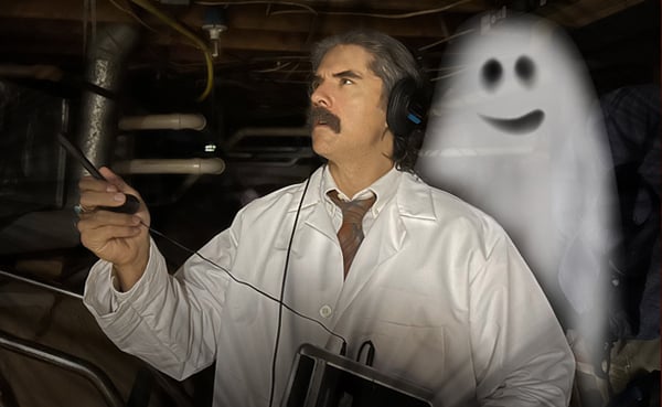 A man in a lab coat and headphones uses a device in a dimly lit room, capturing ghost voices. A cartoon-like spirit appears beside him.