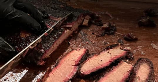 Sliced smoked brisket rests on a wooden cutting board, with a gloved hand deftly holding a knife—perfect for your weekend BBQ.