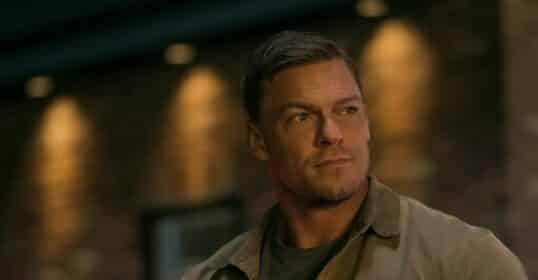 A man in a brown jacket and gray shirt stands in front of a brick wall with dim lighting.