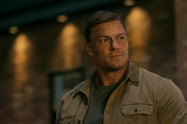 A man in a brown jacket and gray shirt stands in front of a brick wall with dim lighting.
