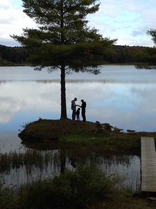 Three people, including a great father-in-law, stand near a tree by the lake. Nearby, a wooden dock stretches out over the water, with empty chairs waiting to host laughter and stories.