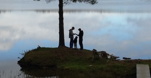 Three people, including the great father-in-law, stand under a tree by a lake, with two benches nearby.