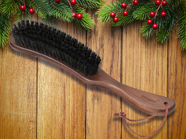 A wooden brush with black bristles and a leather loop rests on a wooden surface, accented by pine branches and red berries above—perfect as unique stocking stuffers for men.