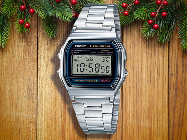 A silver Casio digital wristwatch displays 10:58:50 PM, resting on a wooden surface with evergreen branches and red berries above—an ideal pick for stocking stuffers for men.