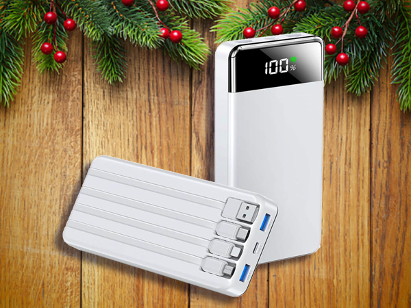 Two white portable power banks with digital displays and built-in charging cables rest on a wooden surface, framed by pine branches and red berries—perfect stocking stuffers for men this holiday season.