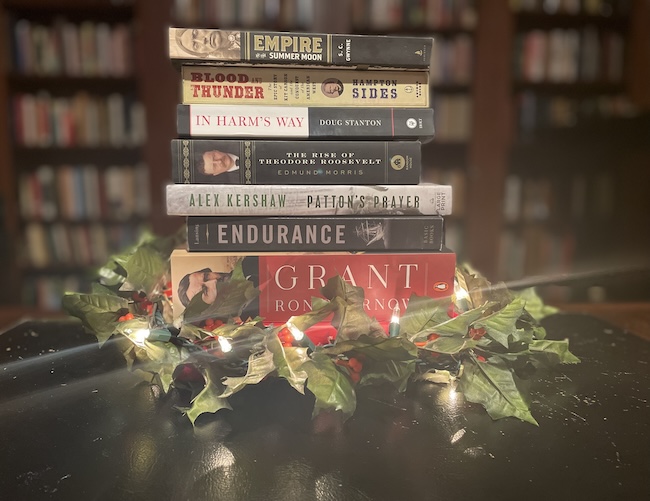 A stack of history books, perfect as a thoughtful Christmas gift, is surrounded by holly and lit with string lights, set against a background of blurred bookshelves.