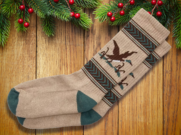 A pair of beige socks with green heels and toes and a bird and tree pattern, perfect as stocking stuffers for men, placed on a wooden surface with pine branches and red berries above.