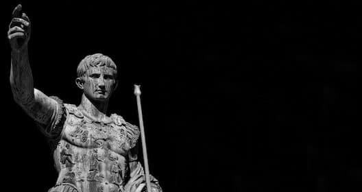 Black and white image of a Roman statue depicting a man in armor holding a staff, with one arm raised against a dark background, as if ready to share heroic tales on an ancient podcast.