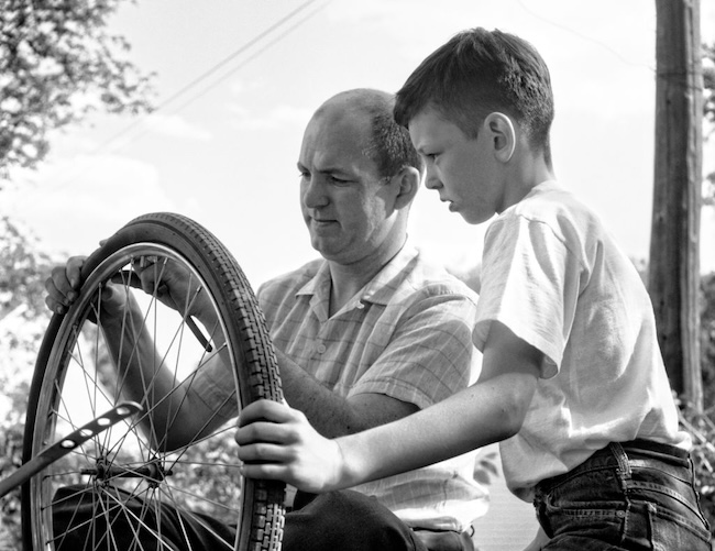 A man and a boy are repairing a bicycle tire outside. Demonstrating the skills of successful fathers, the man carefully explains each step while the boy watches closely, eager to learn.