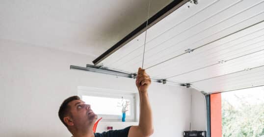 A man pulls the cord of a garage door, opening it partially in a bright garage with shelves and a window, mindful of garage security to prevent potential break-ins.