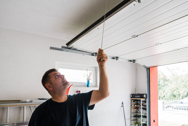 A man pulls the cord of a garage door, opening it partially in a bright garage with shelves and a window, mindful of garage security to prevent potential break-ins.