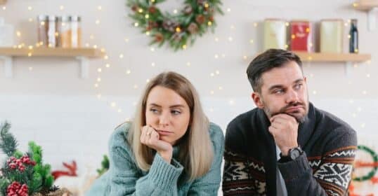 A woman and man lean on a counter, each holding a phone, gazing thoughtfully into the distance. Holiday decorations twinkle in the background, capturing the important moments of togetherness and reflection during this festive season.