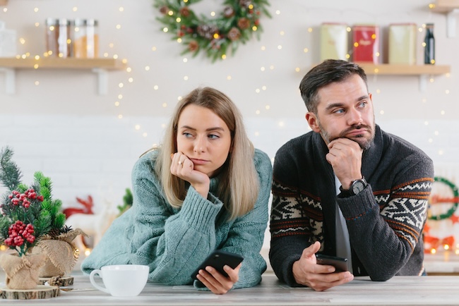 A woman and man lean on a counter, each holding a phone, gazing thoughtfully into the distance. Holiday decorations twinkle in the background, capturing the important moments of togetherness and reflection during this festive season.