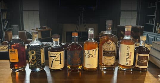 Eight bottles of alcohol-free spirits, including the best zero-proof whiskey, are lined up on a wooden table, with various labels and designs visible.