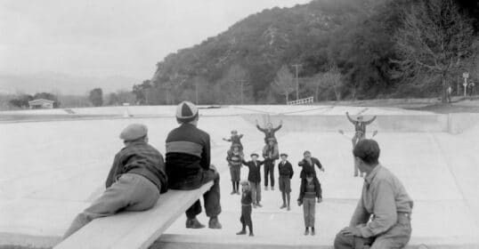 Children play on a seesaw in an outdoor park, their joyful laughter echoing as others stand and cheer. A son eagerly awaits his turn, hoping to join the fun.