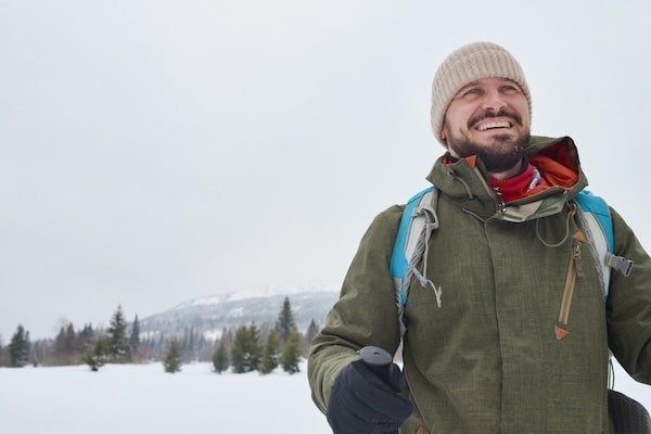 A person in a green jacket and beige beanie is hiking through a winter wonderland, with trees and a majestic mountain in the background.
