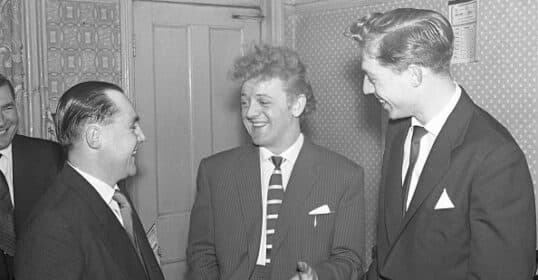 Three men in suits are smiling and conversing in a room with patterned wallpaper, their sense of humor evident. One man holds a cigarette, adding to the laid-back atmosphere where nobody seems to take themselves too seriously.