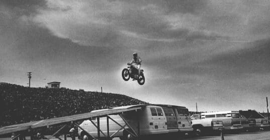 A motorcyclist embodies manliness as he takes the risk to jump over a row of vans on a ramp setup, with a large crowd and a cloudy sky in the background.