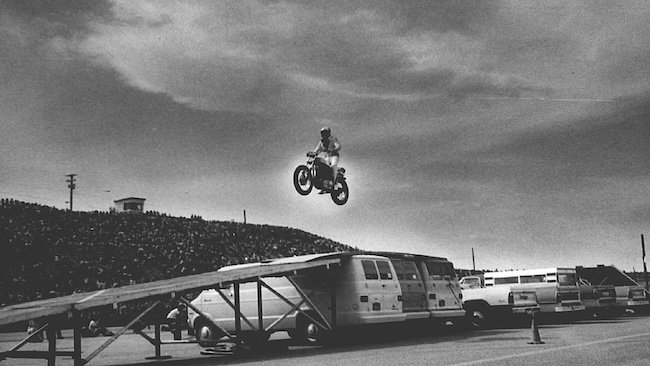 A motorcyclist embodies manliness as he takes the risk to jump over a row of vans on a ramp setup, with a large crowd and a cloudy sky in the background.