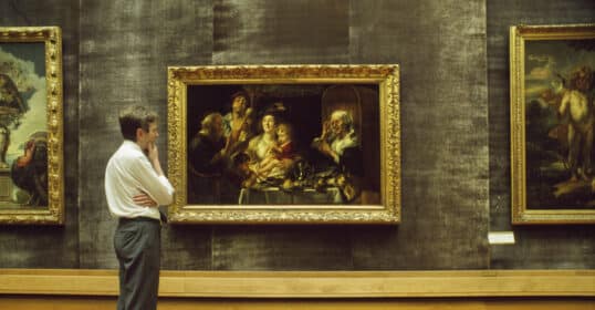 A museum guide stands observing a painting in a gold frame. The artwork depicts a family scene with rich colors.
