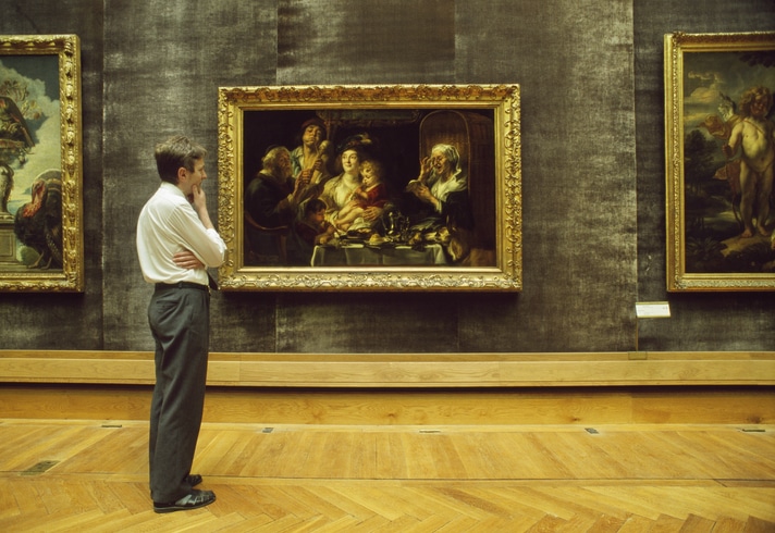 A museum guide stands observing a painting in a gold frame. The artwork depicts a family scene with rich colors.