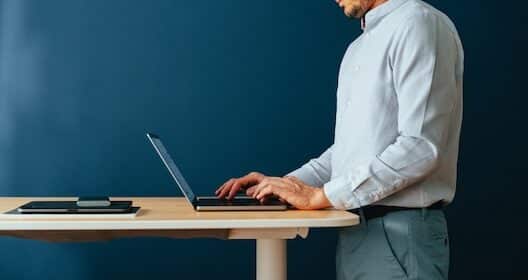 Person in a light blue shirt standing and typing on a laptop at a standing desk against a dark blue background.