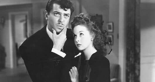 A man with a thoughtful expression holds his chin, contemplating if he's falling in love, while a woman gazes up at him, both dressed in 1940s attire, captured in a classic black and white photograph.