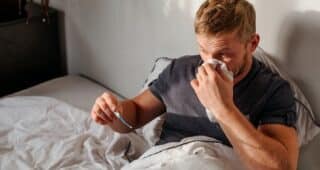 A man is sitting in bed with a thermometer in one hand and a tissue to his nose in the other, battling what feels like a real man cold.