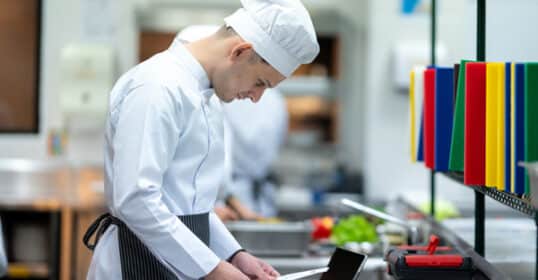In a bustling commercial kitchen, a chef in a crisp white uniform examines a document—an embodiment of his secret to a productive and organized life. Nearby, colorful cutting boards rest neatly on a rack, exemplifying the harmony of mise en place.