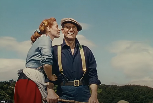 A woman whispers to a smiling man wearing a beret and suspenders, reminiscent of "The Quiet Man," in an outdoor setting under a clear blue sky.