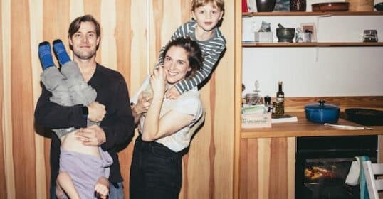 A family of four in a lively kitchen scene showcases their unique parenting style. The father playfully holds an upside-down child, while the mother, embracing another parenting moment, stands with a child joyfully perched on her back.