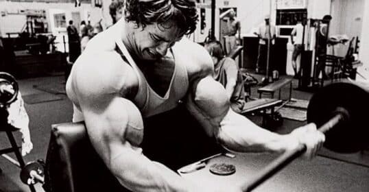 A person in a gym intensely performs a bicep curl with a barbell on a preacher bench. Other gym-goers and equipment are visible in the background.