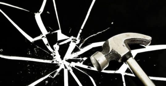 A hammer lies poised beside a radial pattern of broken glass shards on a black background, symbolizing resilience amidst chaos.