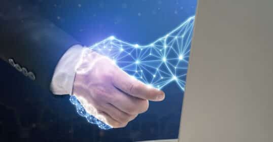 Digital handshake: A glowing wireframe hand emerges from a laptop screen, shaking hands with a human hand, symbolizing AI-driven connection and co-intelligence.