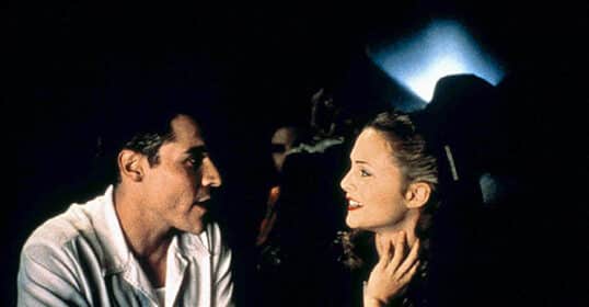 A man in a white shirt talks to a woman in dark clothing at a dimly lit bar; both appear engaged, exchanging classic 1995 flirting tips as they navigate how to flirt in the nostalgic nightlife setting.