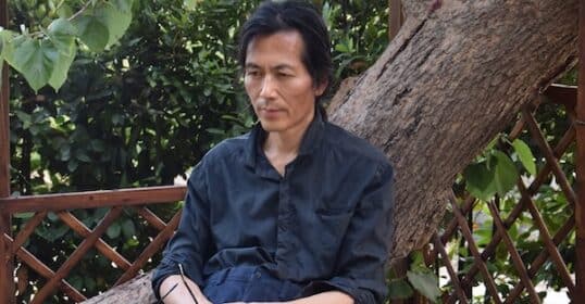 A man in a dark shirt sits on a large tree branch, holding glasses in one hand, with greenery and a wooden lattice in the background.