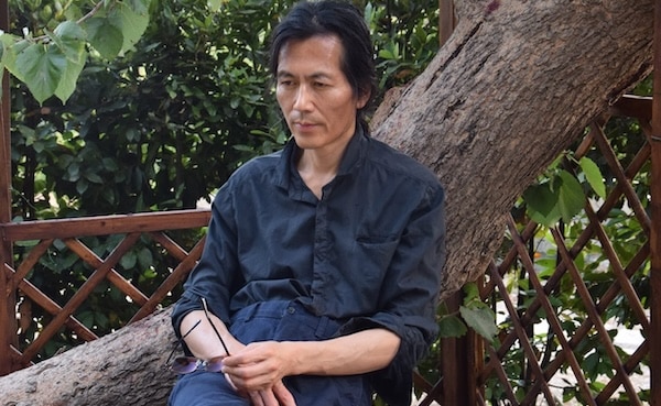 A man in a dark shirt sits on a large tree branch, holding glasses in one hand, with greenery and a wooden lattice in the background.