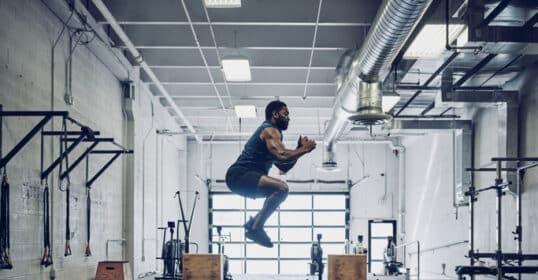 A man showcases his explosive strength with a box jump in the gym, where he's surrounded by weights, exercise balls, and other fitness equipment. Clad in athletic gear, he defies powerpenia with every leap.