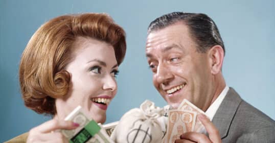 A smiling woman and man hold a bag with a dollar sign and bundles of paper money, appearing pleased and excited as they celebrate reaching personal finance goals in your 40s.