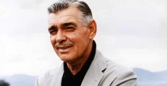 Clark Gable with gray hair and a mustache stands outdoors with arms crossed, showcasing men's fashion in a light gray suit jacket, black shirt, and red pocket square. A rural landscape and mountains are in the background.