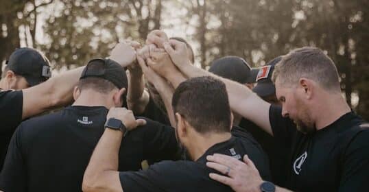 A group of men in black shirts stand outdoors in a circle, raising their joined hands together in a gesture of unity—building tribe and creating communities of men.